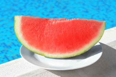 Photo of Slice of fresh juicy watermelon on white plate near swimming pool outdoors, closeup