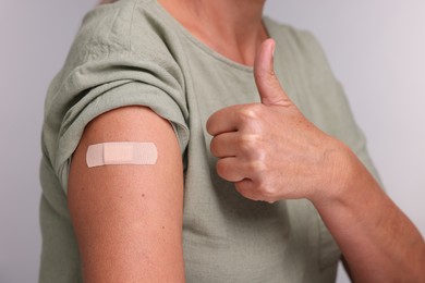 Photo of Woman with adhesive bandage on her arm after vaccination showing thumb up against light grey background, closeup
