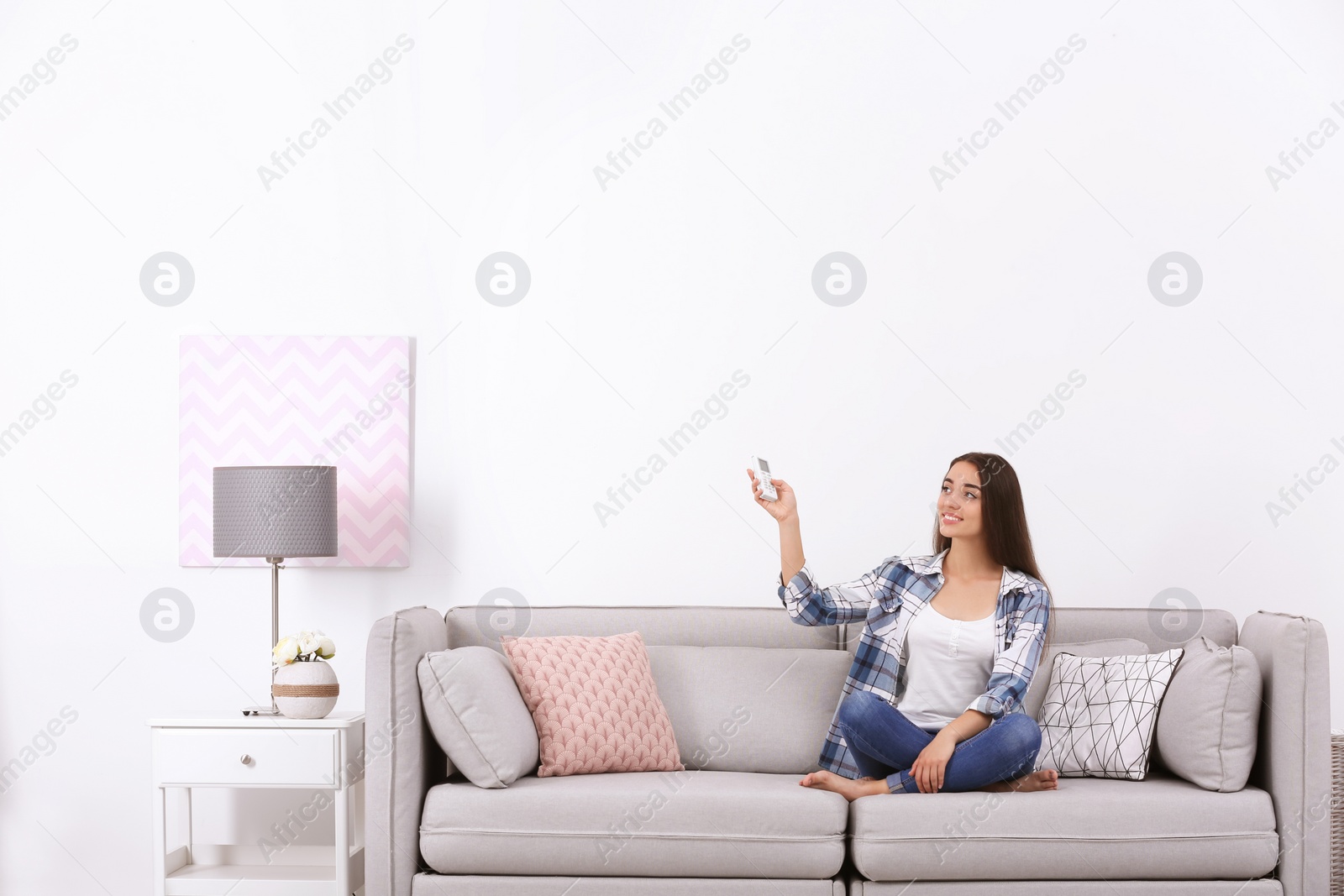 Photo of Young woman switching on air conditioner while sitting on sofa near white wall