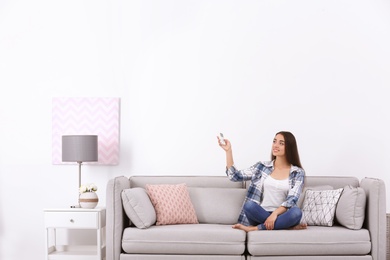 Young woman switching on air conditioner while sitting on sofa near white wall