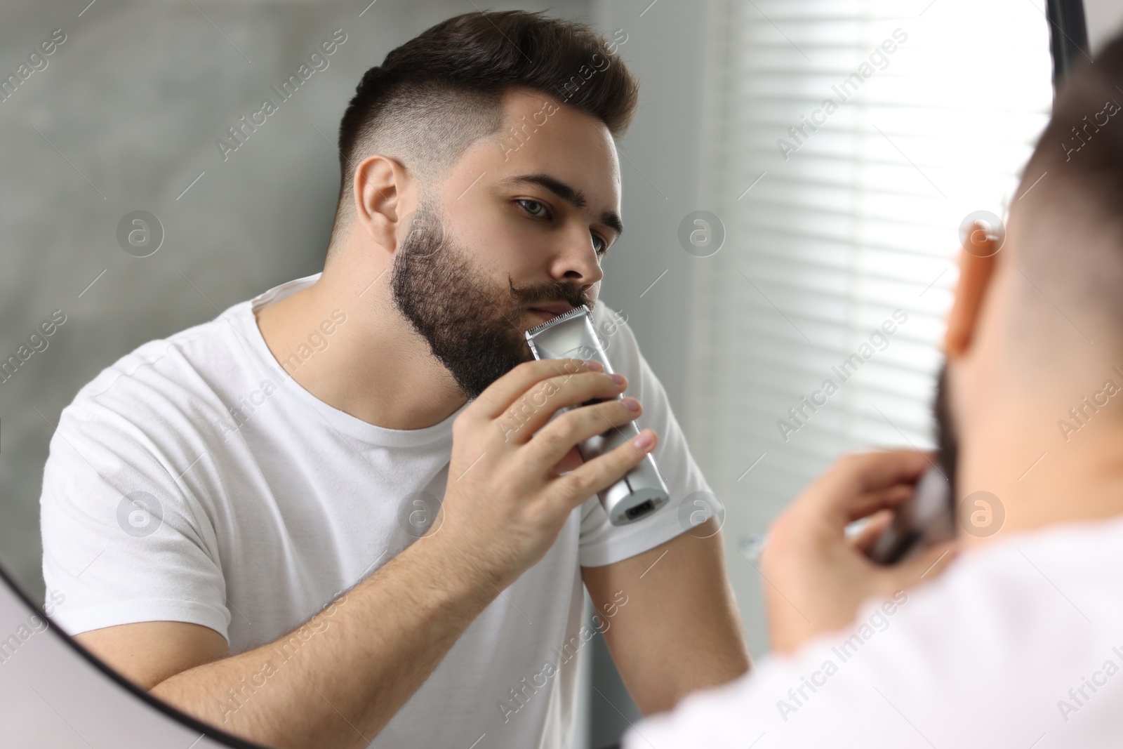 Photo of Handsome young man trimming beard near mirror in bathroom