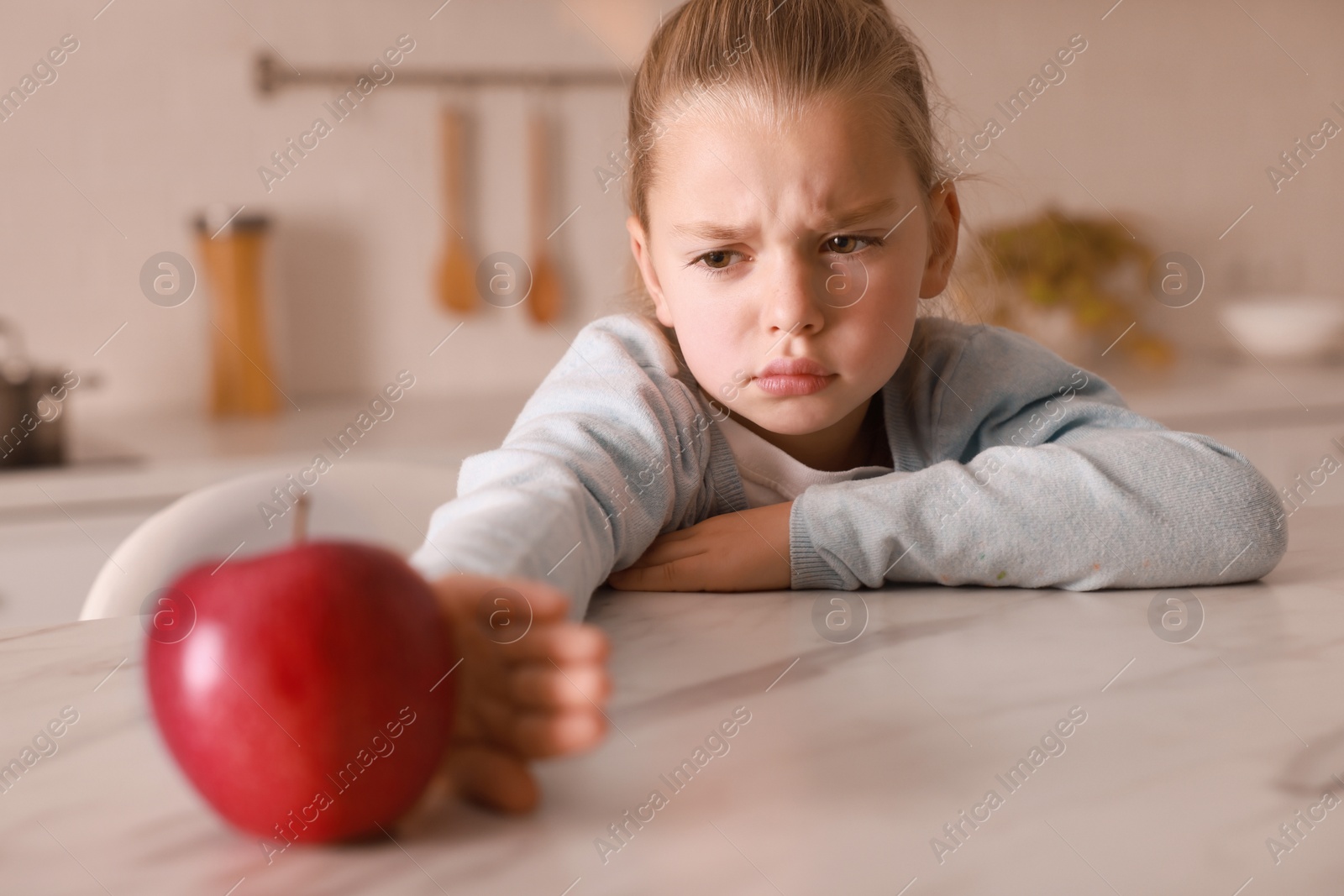 Photo of Cute little girl refusing to eat apple in kitchen