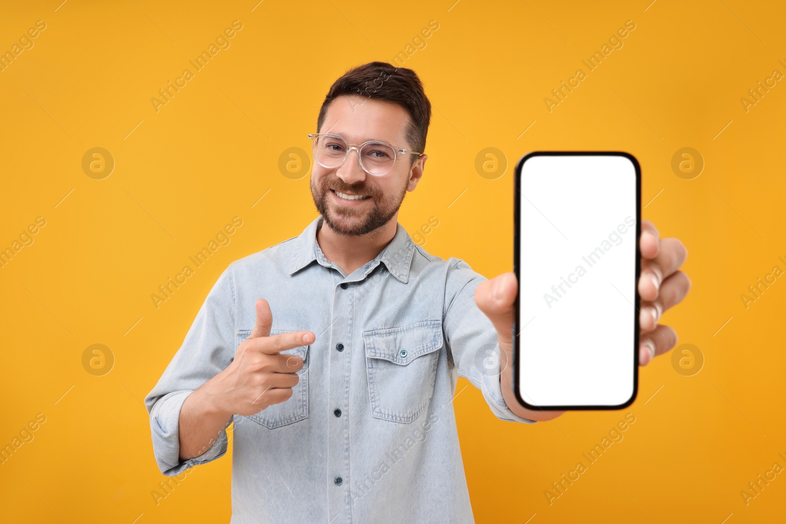 Photo of Handsome man showing smartphone in hand and pointing at it on yellow background