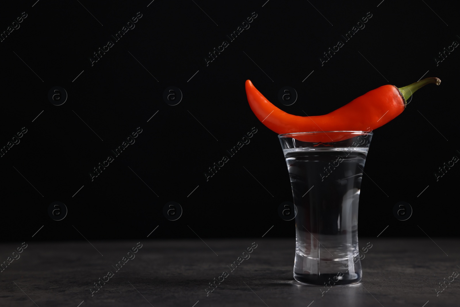 Photo of Red hot chili pepper and vodka in shot glass on grey table against black background, space for text