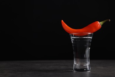 Photo of Red hot chili pepper and vodka in shot glass on grey table against black background, space for text
