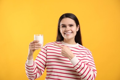 Happy woman with milk mustache pointing at glass of tasty dairy drink on yellow background