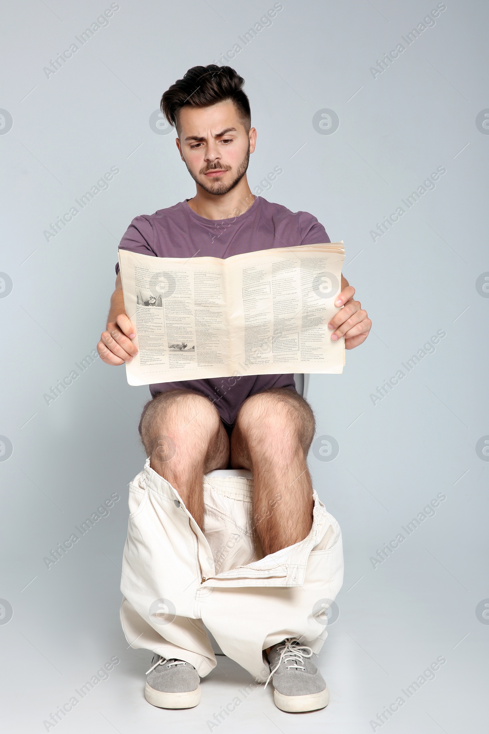 Photo of Young man reading newspaper while sitting on toilet bowl against gray background