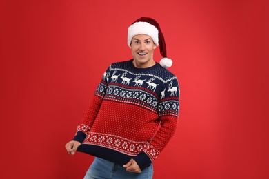 Photo of Happy man in Santa hat showing his Christmas sweater on red background