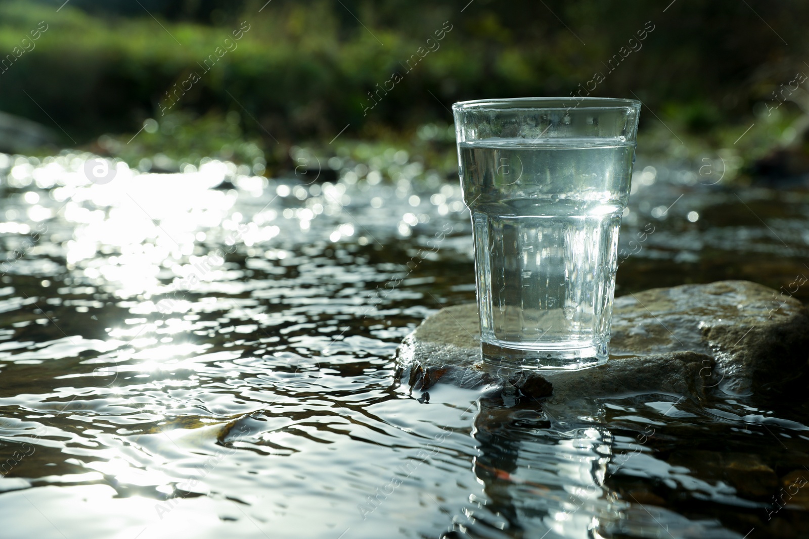 Photo of Glass of fresh water on stone near river outdoors. Space for text