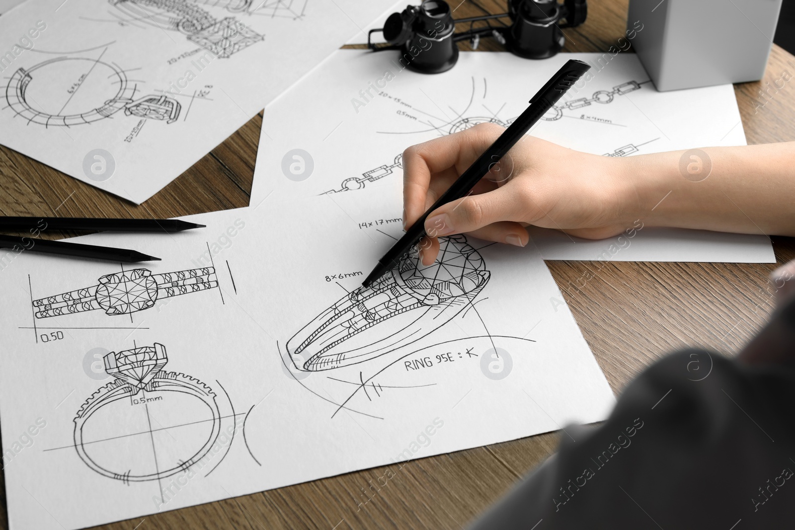 Photo of Jeweler drawing sketch of elegant ring on paper at wooden table, closeup