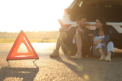 Stressed couple sitting near broken car and warning triangle on roadside