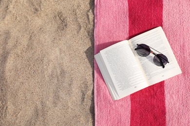 Beach towel with open book and sunglasses on sand, flat lay. Space for text