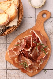 Photo of Slices of tasty cured ham, bread and rosemary on white tiled table, flat lay