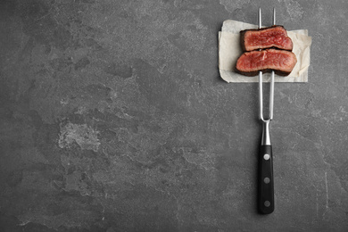 Photo of Carving fork with slices of delicious beef steak on grey table, top view. Space for text