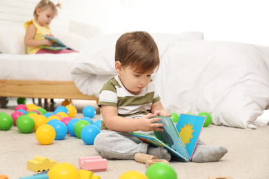 Photo of Cute little boy with book playing on floor while girl sitting on bed. Children's development