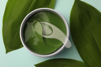 Photo of Jar of under eye patches with spoon and green leaves on turquoise background, flat lay. Cosmetic product