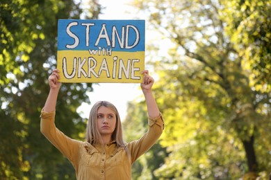 Sad woman holding poster in colors of national flag and words Stand with Ukraine outdoors. Space for text