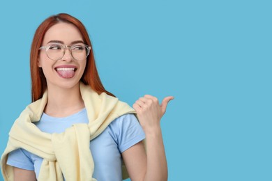 Photo of Happy woman showing her tongue and pointing at something on light blue background. Space for text