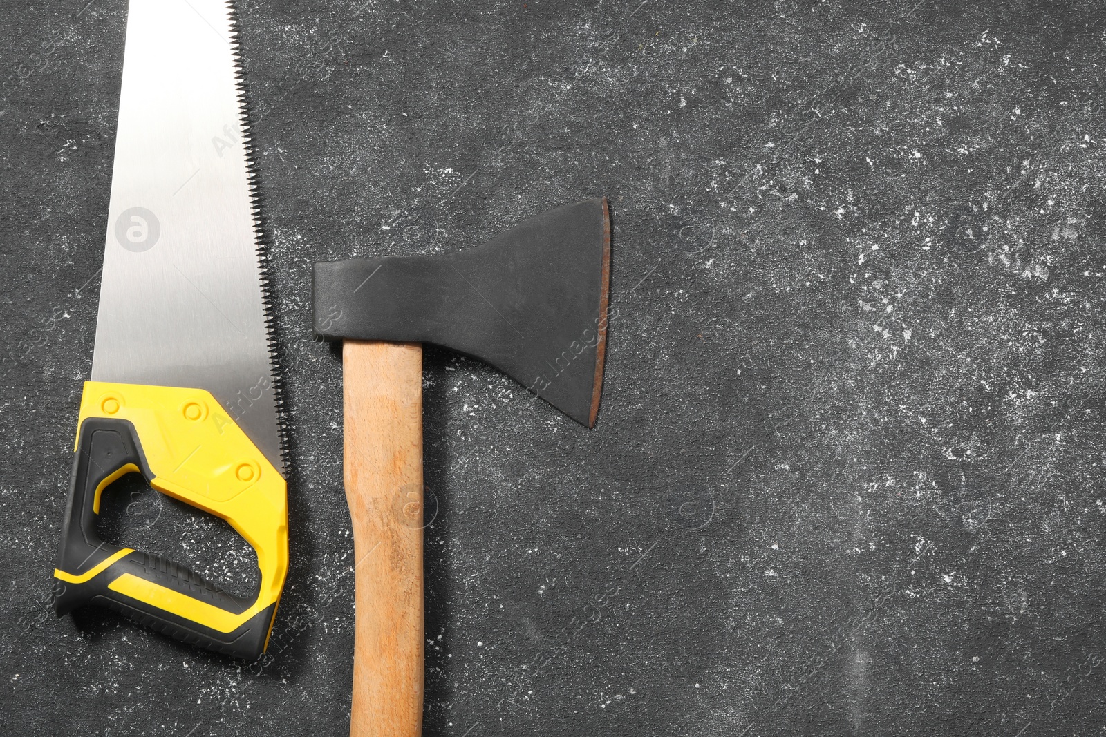 Photo of Saw with yellow handle and axe on dark gray textured background, flat lay. Space for text