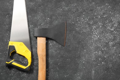Photo of Saw with yellow handle and axe on dark gray textured background, flat lay. Space for text