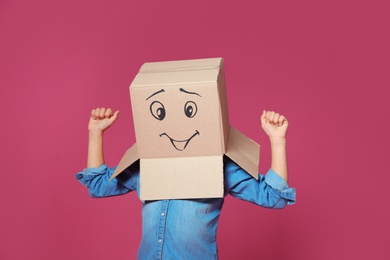 Photo of Cute little child wearing cardboard box with smiling face on color background