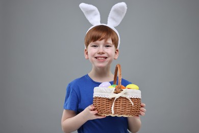Easter celebration. Cute little boy with bunny ears and wicker basket full of painted eggs on grey background