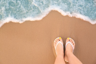 Photo of Top view of woman with white flip flops on sand near sea, space for text. Beach accessories