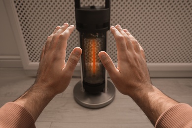 Photo of Man warming hands near modern heater indoors, closeup