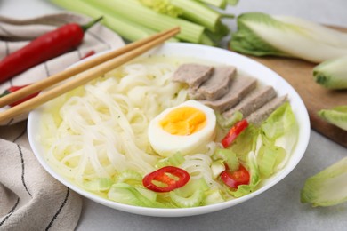 Photo of Bowl of delicious rice noodle soup with meat and egg on light grey table, closeup