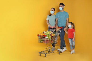 Family with protective masks and shopping cart full of groceries on yellow background. Space for text