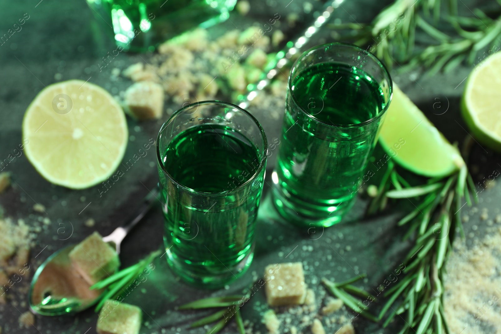 Photo of Absinthe in shot glasses, spoon, brown sugar, lime and rosemary on gray table, closeup. Alcoholic drink