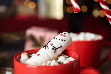 Funny marshmallow snowman in cup of hot drink against blurred festive lights, closeup