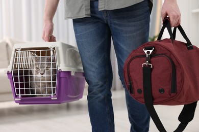 Photo of Travel with pet. Man holding carrier with cute cat and bag at home, closeup