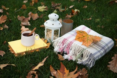 Composition with cup of coffee, book, plaid and autumn leaves on green grass outdoors