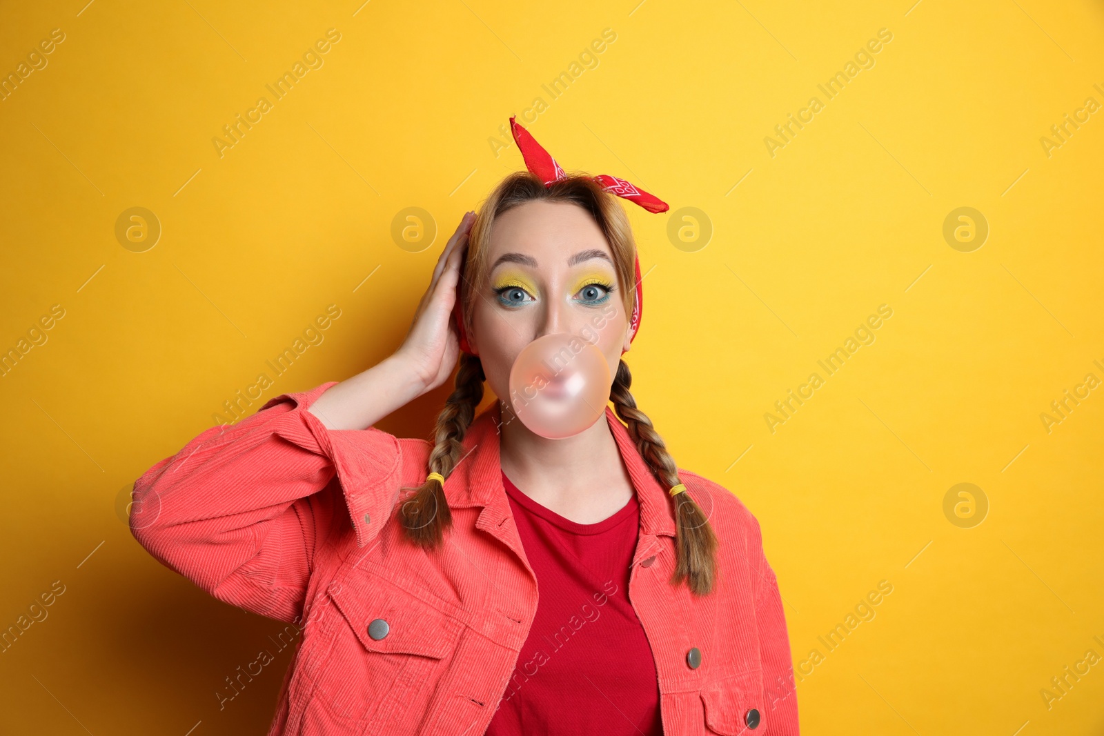 Photo of Fashionable young woman with braids and bright makeup blowing bubblegum on yellow background