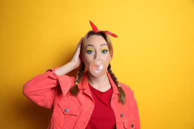 Fashionable young woman with braids and bright makeup blowing bubblegum on yellow background