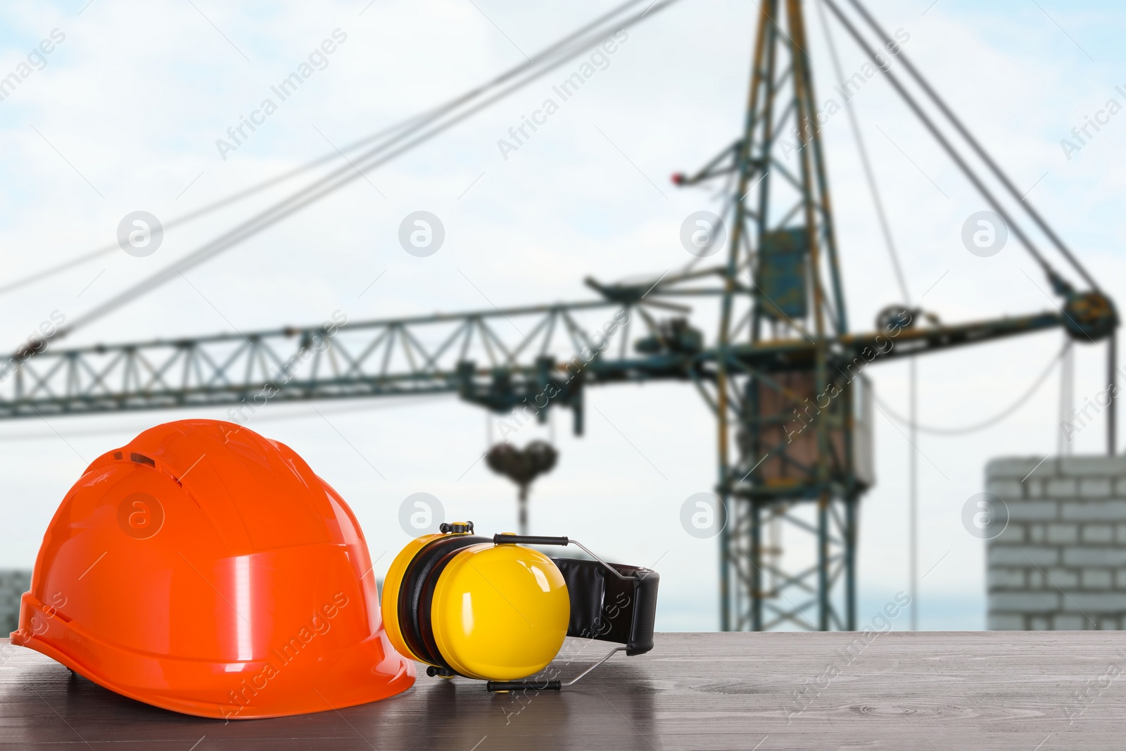 Image of Safety equipment. Hard hat and protective headphones on wooden surface near construction site with tower crane outdoors, space for text