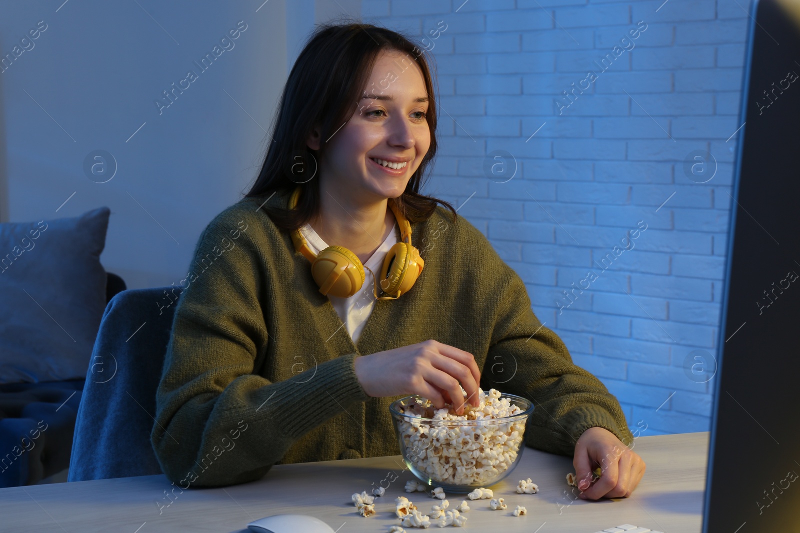 Photo of Beautiful young woman with headphones and popcorn watching film on computer at table indoors at night