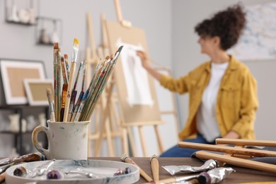 Photo of Woman painting on easel with paper in studio, focus on brushes