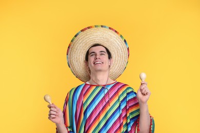 Young man in Mexican sombrero hat and poncho with maracas on yellow background