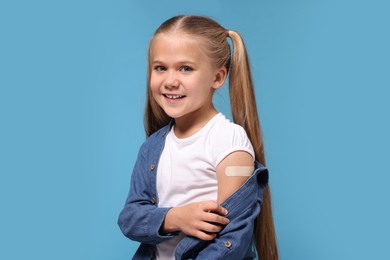 Photo of Happy girl with sticking plaster on arm after vaccination against light blue background