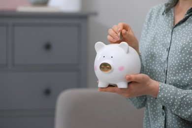 Photo of Young woman putting coin into piggy bank indoors, closeup