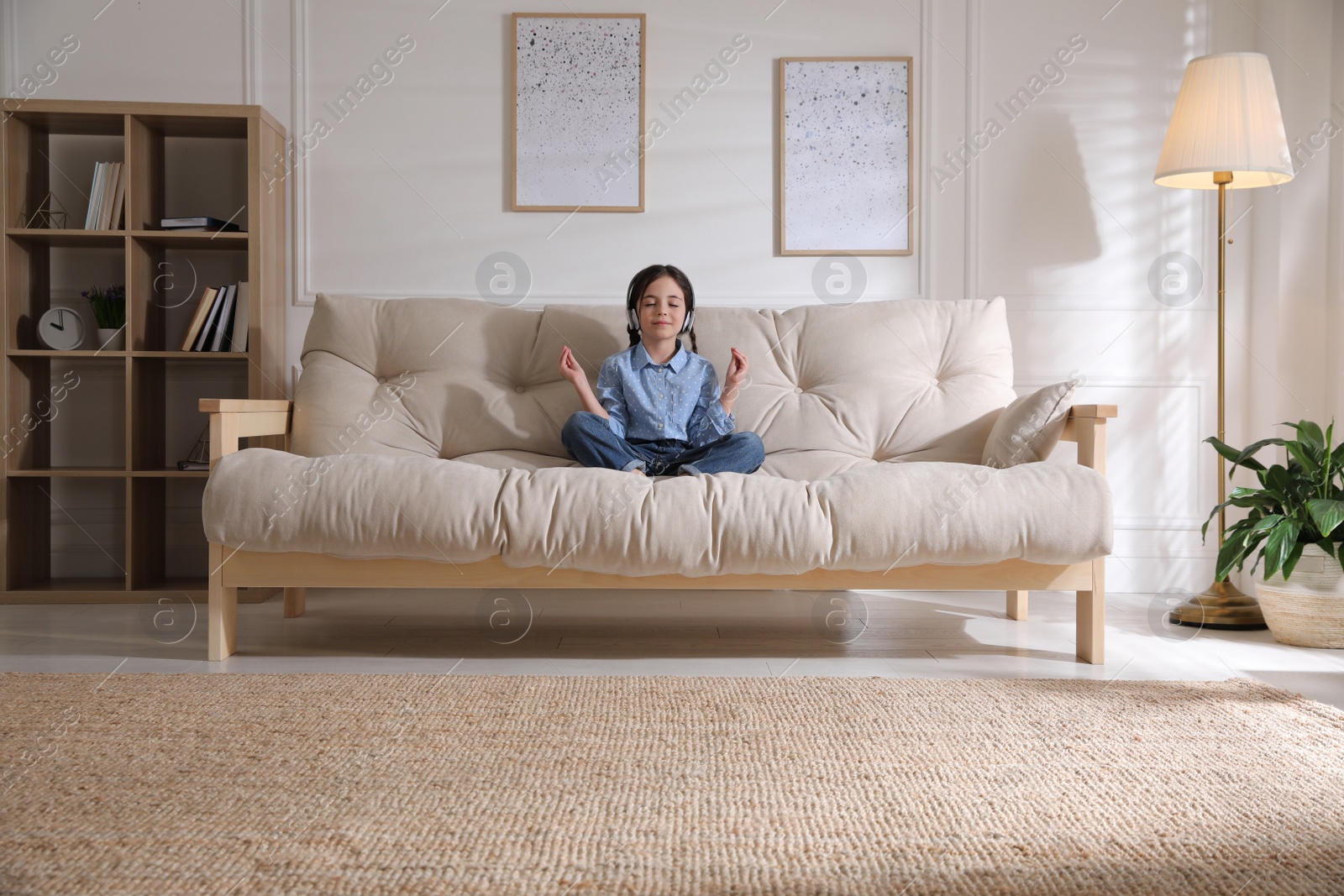 Photo of Little girl with headphones meditating on sofa at home