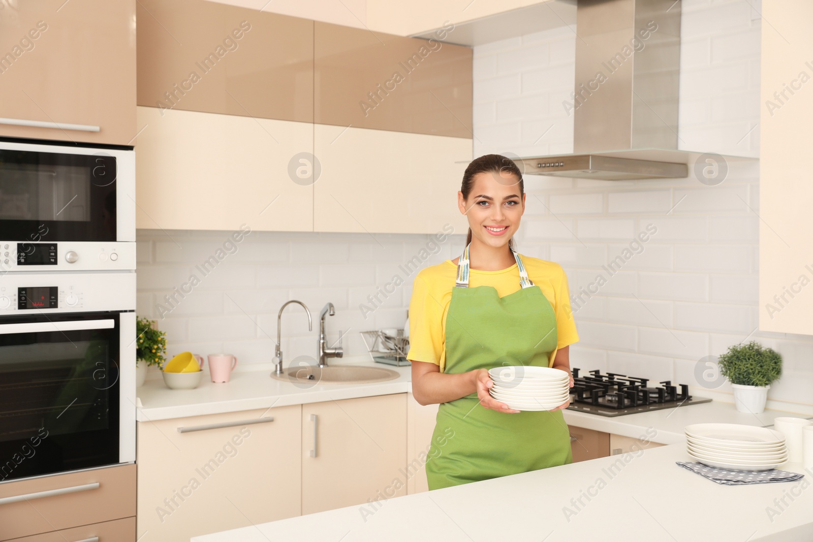 Photo of Young woman holding stack of clean dishes in kitchen