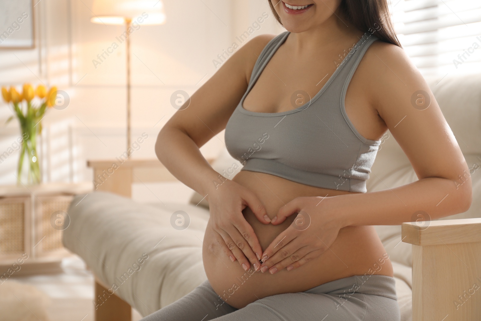 Photo of Pregnant young woman making heart with her hands on belly at home, closeup. Space for text