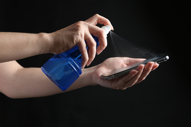 Photo of Woman spraying antiseptic onto smartphone against black background, closeup