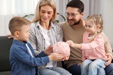 Photo of Family budget. Little girl putting coin into piggy bank while her parents and brother watching indoors
