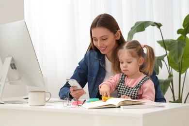 Photo of Woman working remotely at home. Mother using phone while her daughter playing at desk
