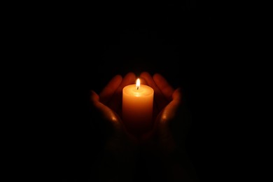 Woman holding burning candle in hands on black background, closeup