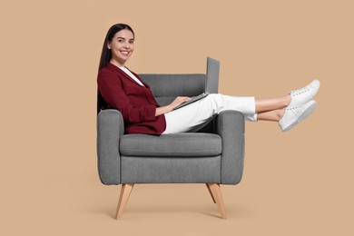 Happy woman with laptop sitting in armchair on beige background